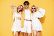 © halayalex - Group of young three stylish friends posing in studio. Fashion man and two cute female dressed in casual summer clothes. Smiling models having fun. Cheerful women and guy near yellow wall, sunglasses
