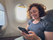© Юлия Завалишина - Passenger woman is flying in plane. Girl using phone sitting by airplane window. Traveling female with smartphone inside plane enjoying flight. Woman using wifi connection on board. Traveling girl
