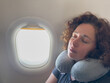 © Юлия Завалишина - Rest on plane. Young woman napping sitting by porthole in business jet using neck pillow for travel. Passenger girl flying in plane. Girl sleeping in airplane going on trip vacation. Traveling girl