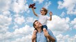 © zephyr - Happy father carries his joyful little son on his shoulders as the young boy holds a small red toy airplane up toward the blue sky