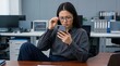 © illustratica - Young East Asian woman wearing glasses squints at her smartphone screen in an office