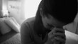 © Marco - Close-up of woman sitting with head bowed and hands clasped, expressing emotional exhaustion, reflection, and quiet despair in intimate home setting, black and white