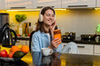 © Andrii Iemelianenko - Young woman smiling in kitchen wearing headphones streaming favorite music on smartphone during break. Adult girl enjoying calm leisure at home with coffee playlist focus for stress relief and mood.