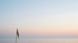 © byvea - A solitary, weathered flag stands against the vast, serene expanse of pastel sky merging with the tranquil ocean horizon, evoking profound peace and solitude