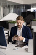© fizkes - Focused businessman in suit sit at desk in contemporary office, using smartphone for work communication, multitasking with digital devices for efficient business management and professional networking