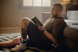 © Seventyfour - Young man reading book at home while wearing electronic ankle monitor, representing house arrest and criminal justice surveillance. Useful for legal, prison reform, probation, rehabilitation campaigns