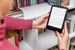 © Oleg Opryshko - a woman holds an e-book in her hand. A woman reads an e-reader near a bookcase. an e-book against the background of paper books. ease of use of an e-book. a modern way to read books