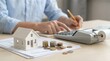 © Mariya Sorvacheva - Person calculating rent payments with a calculator, stacks of coins, and a small house model on financial documents on a wooden desk in a bright indoor setting