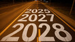 © Zara - Road markings indicate the years twenty twenty-five, twenty twenty-seven, and twenty twenty-eight painted in white on the dark asphalt surface of an empty highway during the late night hours.
