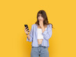 © Designerant - Worried young woman looking at smartphone and biting nail with anxious expression, reacting to problem or message concept. Isolated on bright yellow background with copy space for advertising use.