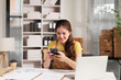 © MINAE - Young Asian woman entrepreneur working at a home office, handling her online business, using a smartphone for communication and order management, with a laptop and documents on the desk
