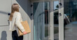 © valiantsin - Woman carrying cardboard box at modern entrance. She waits by glass doors in bright daylight. Moving home delivery and relocation concept..