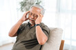 © chokniti - Elderly man with headache and eye strain sitting on sofa in bright home interior