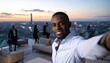 © slametamirin - Smiling man taking selfie on rooftop with city view at sunset.