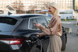 © vladim_ka - Driver operating charging process for black electric vehicle at urban charging station. The individual is engaged with a smartphone while connecting the charger, dressed in casual professional attire.