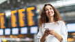© Bonsales - Young woman smiling, standing in an airport terminal, holding a smartphone, and looking up at a departure board