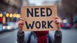 © Visglow - A person holding a Need Work sign is seen in the city, illustrating a call for employment.