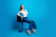 © deagreez - Happy young woman posing on a modern stool against a bright blue background in casual jeans and heels, stylish portrait conveying confidence, relaxation and contemporary fashion