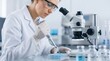 © Dreamscape - Female scientist in white coat and safety glasses using micropipette to transfer liquid sample into multiwell plate under microscope for laboratory research, biotechnology