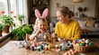© horizon - Smiling young mother in a yellow sweater and her little daughter wearing pink bunny ears joyfully paint Easter eggs at a dining room table.