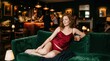 © Fiverr/fazilazia703 - Elegant woman in red silk lingerie relaxing on a green velvet sofa, enjoying a cocktail in a sophisticated, dimly lit bar with a bustling background.