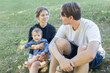 © Anna - happy parents chat while walking with baby girl in park, sitting on grass. loving mom, dad with their 6-month-old daughter enjoy time spent together. Family Day. tender moment of parental interaction