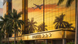 © hamad - A grand luxury hotel building with a golden glass facade beautifully reflects palm trees, a sunset sky, and an airplane overhead.