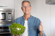 © luciano - Smiling mature man holding a bowl of fresh lettuce looking at camera with a thumbs-up, cooking healthy food at home in a modern kitchen, preparing vegetables. Concept of nutrition and healthy eating
