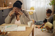 © Seventyfour - African American mother covering face while reviewing household bills, struggling with financial stress as children playing nearby. Ideal for poverty, debt, budgeting, family hardship themes