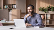 © insta_photos - Smiling bearded indian businessman working on laptop in modern office lobby space. Young indian student using computer remote studying, watching online webinar, virtual training on video call meeting.
