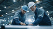 © sam richter - Two experienced engineers wearing hard hats examine technical blueprints inside a large industrial facility.