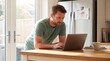 © ryker - Man leaning on kitchen island working on laptop, focused expression, natural morning light and family drawing on refrigerator conveying home office routine and calm productivity