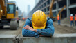 © miss irine - Construction worker in yellow hard hat rests head on arms on concrete block. Exhausted man on building site. Machines and workers in background.
