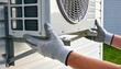 © Netsai - Gloved hands carefully positioning a white outdoor air conditioning condenser unit onto a metal wall bracket.