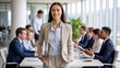 © Lamia - Confident young businesswoman standing in a modern office boardroom, smiling at the camera while her diverse team collaborates