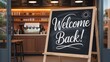 © EmonKumar - Welcome back sign on a chalkboard easel at the entrance of a cozy modern cafe or coffee shop.