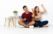 © luismolinero - Young couple sitting on the floor isolated on white background celebrating a victory
