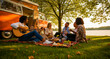 © Fandom - Friends having a picnic and playing guitar by a vintage camper van near a lake. Group laughing and drinking wine under a tree at sunset. Summer travel and leisure concept for lifestyle marketing.