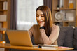 © 220 Selfmade studio - Happy Asian woman looking laptop computer smiling relaxed resting hand chin sitting desk comfortable living room working from home enjoying online entertainment or video call with friends