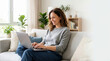 © Xenia - Smiling woman sitting on a couch and using a laptop in a bright living room. Happy female freelancer working remotely or browsing the internet at home. Comfortable domestic lifestyle