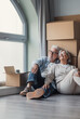 © Daniel - A happy senior couple relaxes on the floor beside packed moving boxes, smiling and looking out a window as they settle into their new home during retirement and relocation.
