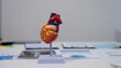 © NanSan - Anatomical human heart model on a stand on a desk with blurred medical charts and documents in the background, healthcare education and cardiology concept.