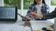 © NanSan - Architects reviewing house floor plans at desk, laptop and tablet in bright office, collaborating on home design project with blueprints and documents.