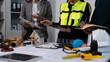 © NanSan - Team meeting with engineer in safety vest reviewing plans at desk with scale model house, hard hat, tools and documents; architecture, construction and project management concept.