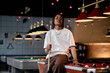© TrueFrame Collective - Portrait of young adult Black man holding pool cue while sitting on billiards table, representing leisure venue culture and social entertainment. Useful for nightlife marketing and youth lifestyle cam