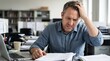 © haris - A stressed middle-aged man sitting at his desk in an office, holding his head in his hands and looking at papers.