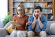 © Liubomir - Young couple sitting on a sofa at home, feeling stressed and anxious after opening an important letter containing bad news about finances, bills, or debt