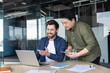 © Liubomir - Two diverse male colleagues collaborating on a laptop, discussing business ideas and sharing information, fostering teamwork and innovation in a contemporary office workspace