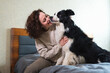 © Юлия Завалишина - Smiling woman playing with cute puppy dog border collie on bed at home indoor. Owner girl stroking holding dog friend sitting on sofa in living room. Love for pets friendship support team concept