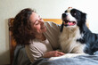 © Юлия Завалишина - Smiling woman playing with cute puppy dog border collie on bed at home indoor. Owner girl stroking holding dog friend sitting on sofa in living room. Love for pets friendship support team concept